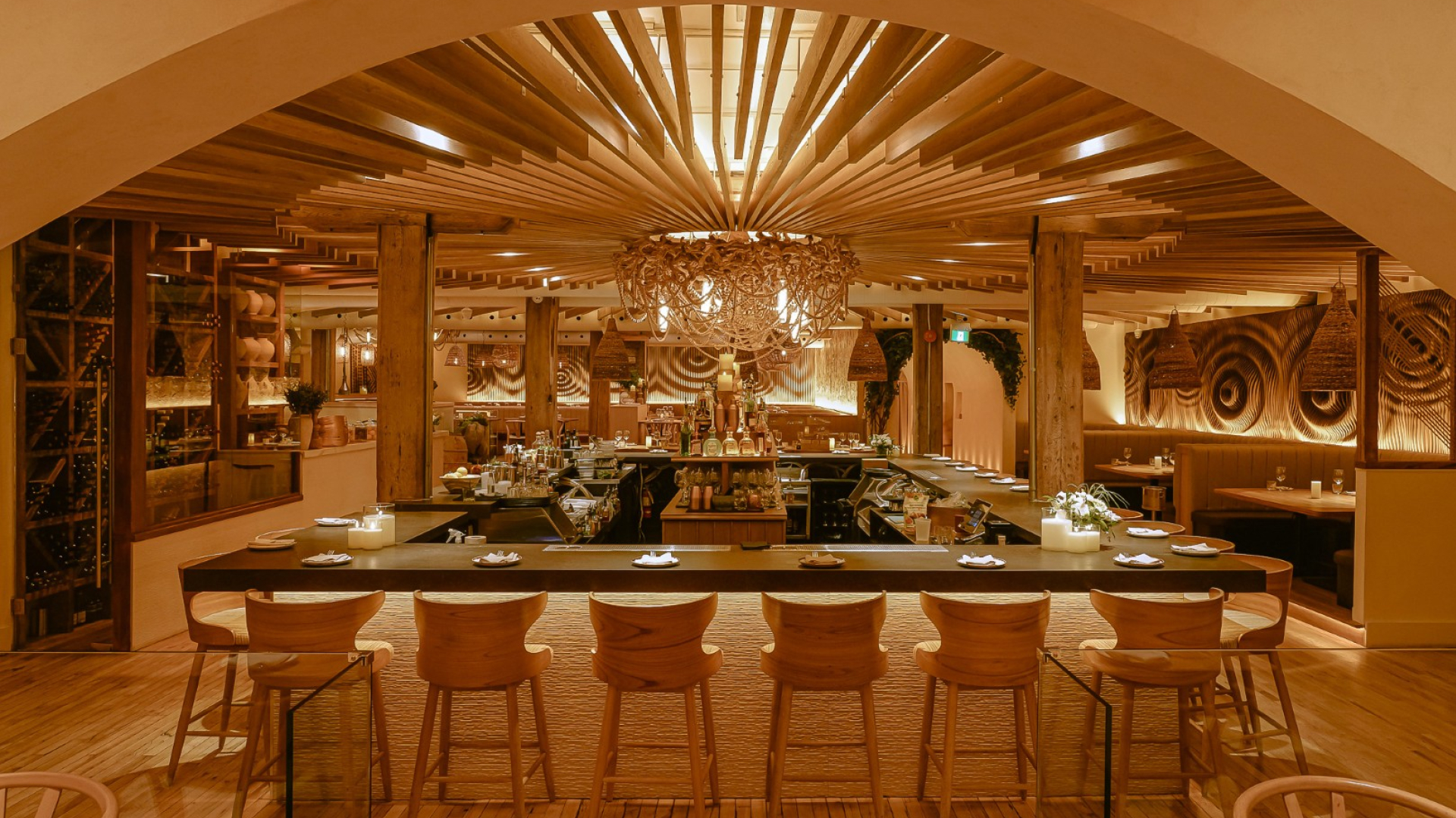 A warmly lit restaurant interior with a long central bar and wood stools facing an open kitchen. Curved arches, layered wood ceiling details, and decorative lighting create an intimate, upscale dining atmosphere.